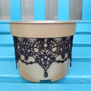 May include: A gold-coloured plant pot with a black lace band around the middle. The lace has an intricate floral design with teardrop-shaped pendants. The pot sits on a blue surface.