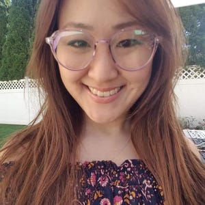 May include: A woman with long brown hair wearing a floral off-the-shoulder top and clear purple-tinted glasses. She is smiling at the camera.