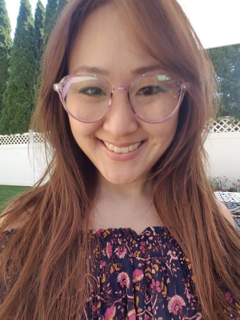 May include: A woman with long brown hair wearing a floral off-the-shoulder top and clear purple-tinted glasses. She is smiling at the camera.