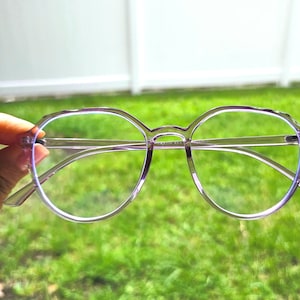 May include: Purple round-framed glasses with clear lenses. The glasses are held in a hand against a green grassy background.