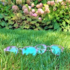 May include: Three pairs of pink-rimmed glasses with blue lenses lie in a bed of green grass. The glasses are partially obscured by the grass.