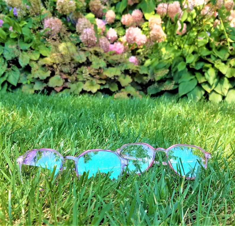 May include: Three pairs of pink-rimmed glasses with blue lenses lie in a bed of green grass. The glasses are partially obscured by the grass.