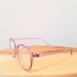 May include: Pink plastic framed glasses with clear lenses. The glasses are on a wooden surface.