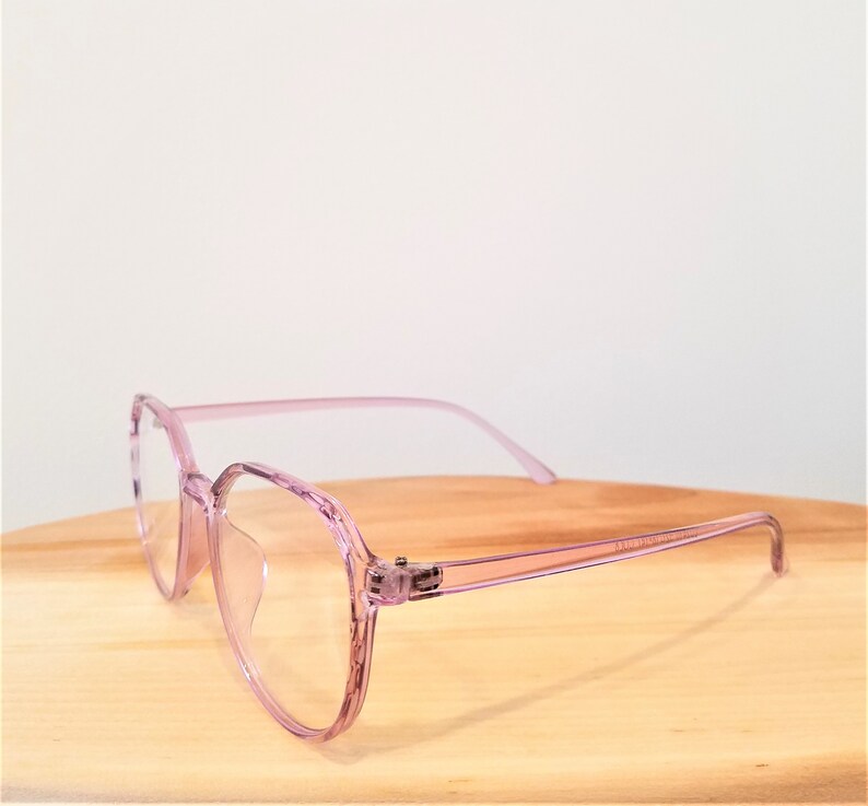 May include: Pink plastic framed glasses with clear lenses. The glasses are on a wooden surface.