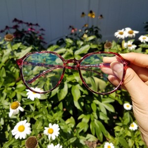 以下が含まれることがあります： 赤と透明な丸い眼鏡で、べっ甲模様が施されています。眼鏡は白い花と緑の葉の背景の前に置かれています。