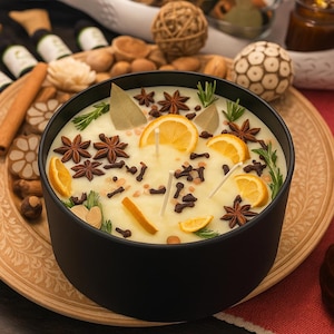 May include: A large, round candle in a black bowl, decorated with orange slices, star anise, cloves, and rosemary. The candle sits on a wooden tray, with other decorative items in the background.