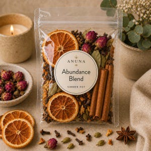 May include: A clear plastic bag filled with a simmer pot blend, featuring dried orange slices, cinnamon sticks, rosebuds, and other spices. The label reads "Anuna Abundance Blend Simmer Pot". A lit candle and small bowl of rosebuds are in the background.