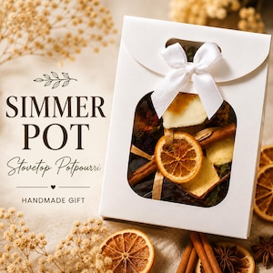 May include: A white gift box with a window, tied with a white ribbon, containing a simmer pot kit. The kit includes dried orange slices, cinnamon sticks, and other botanicals. Text on the box reads "Simmer Pot, Stovetop Potpourri, Handmade Gift."