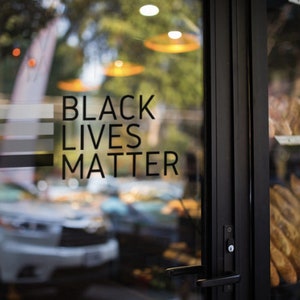 May include: A glass door with a black "Black Lives Matter" sign on it. The sign has five horizontal stripes in shades of gray and black. The door is open and there are loaves of bread visible through the door.