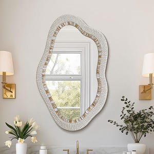 May include: An irregular, organic-shaped wall mirror featuring a textured white frame with subtle gold-toned accents. The mirror has a wavy, non-symmetrical border and is mounted on a wall above a wooden vanity cabinet with a white marble countertop.