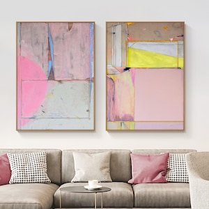 May include: Two framed abstract paintings with pink, yellow, and gray geometric shapes. The artwork hangs above a gray sofa with pink and patterned throw pillows. The paintings have gold frames and are displayed in a living room setting.