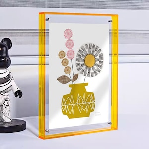 Puede incluir: Un marco de fotos de acrílico amarillo con un passe-partout blanco y una impresión de un arreglo floral estilizado en un jarrón amarillo. La impresión se titula "Flora Botanical".