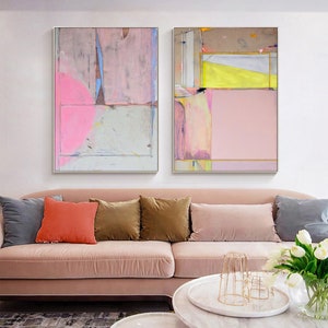 May include: Two framed abstract paintings with pink, yellow, and grey geometric shapes. The artwork hangs above a pink sofa with colourful throw cushions. The paintings have a modern, minimalist style, adding a touch of contemporary art to the living room.