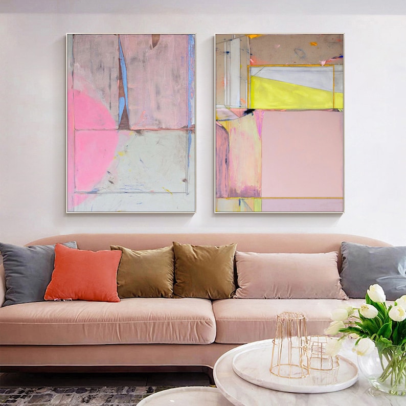 May include: Two framed abstract paintings with pink, yellow, and grey geometric shapes. The artwork hangs above a pink sofa with colourful throw cushions. The paintings have a modern, minimalist style, adding a touch of contemporary art to the living room.