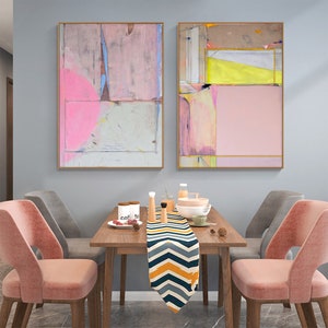 May include: Two abstract paintings in gold frames, featuring pink, grey, and yellow tones. They hang above a wooden dining table with a patterned runner, tableware, and pink velvet chairs.