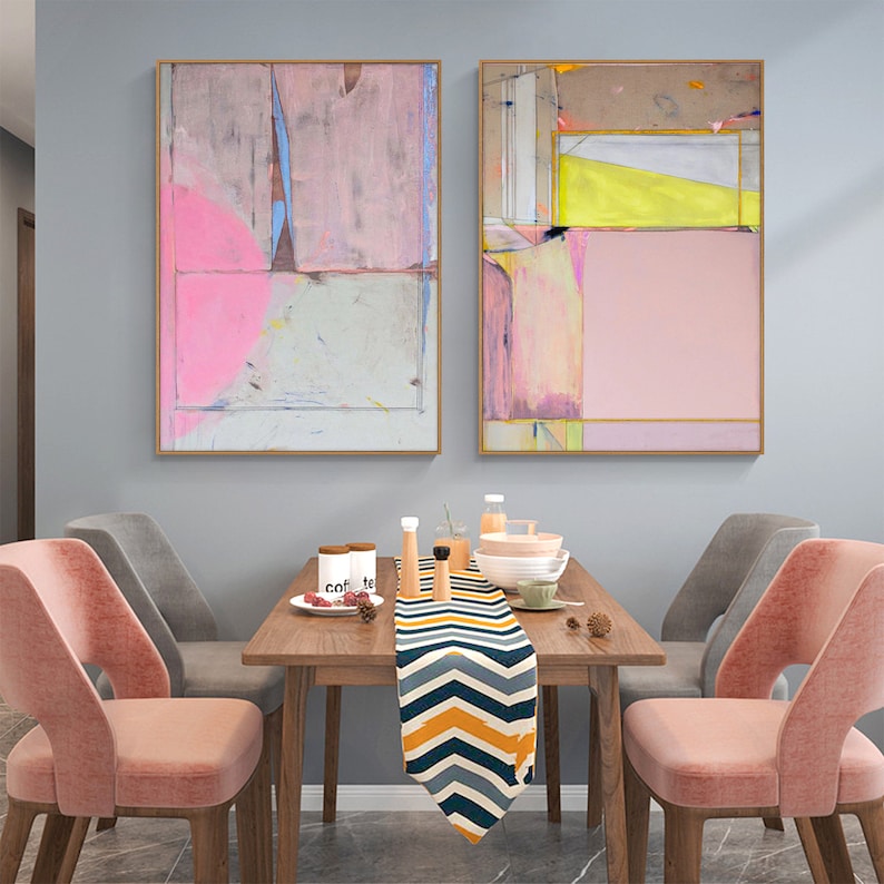 May include: Two abstract paintings in gold frames, featuring pink, grey, and yellow tones. They hang above a wooden dining table with a patterned runner, tableware, and pink velvet chairs.