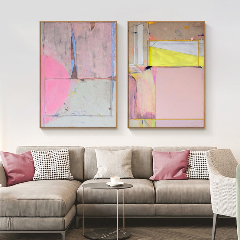 May include: Two framed abstract paintings with pink, yellow, and gray geometric shapes. The artwork hangs above a gray sofa with pink and patterned throw pillows. The paintings have gold frames and are displayed in a living room setting.