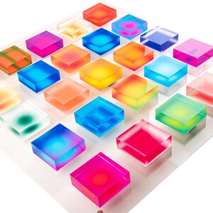 Puede incluir: Una pantalla de acr&iacute;lico transparente con 25 bloques cuadrados transl&uacute;cidos de colores. Los bloques est&aacute;n dispuestos en un patr&oacute;n de cuadr&iacute;cula, mostrando un espectro de colores vibrantes, incluyendo rojo, azul, amarillo y verde. La pantalla est&aacute; montada sobre una base de acr&iacute;lico transparente.