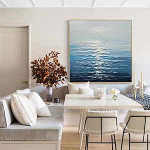 May include: A painting of a seascape with blue and white waves, a light blue sky, and a few small boats in the distance. The painting is hanging on a wall in a room with a white couch, a white table, and two chairs.