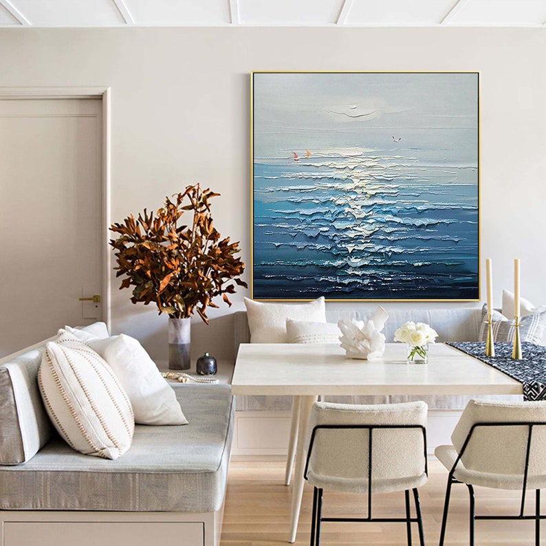 May include: A painting of a seascape with blue and white waves, a light blue sky, and a few small boats in the distance. The painting is hanging on a wall in a room with a white couch, a white table, and two chairs.
