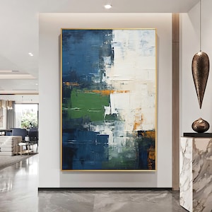 May include: Abstract painting with bold strokes of blue, green, white, and gold paint. The painting is framed in a gold frame and hangs on a white wall.