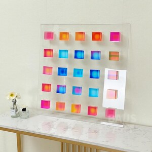 May include: A wall art piece featuring a grid of 25 colorful, translucent cubes. The cubes are arranged in a gradient pattern, transitioning from pink to orange to blue. The cubes are mounted on a clear acrylic panel.
