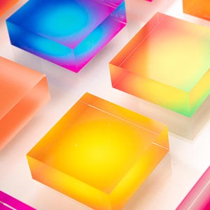 Puede incluir: Una serie de bloques cuadrados transl&uacute;cidos y coloridos. Los bloques muestran un degradado de colores, incluyendo azul, rosa, amarillo y naranja. Los bloques est&aacute;n dispuestos sobre una superficie blanca, creando una exhibici&oacute;n vibrante y llamativa.