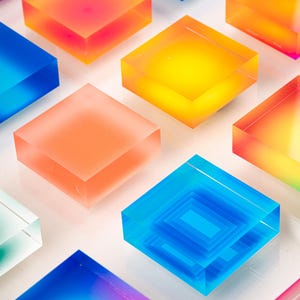 Puede incluir: Una serie de bloques cuadrados transl&uacute;cidos de colores en tonos azules, naranjas, amarillos y rosas. Los bloques est&aacute;n dispuestos sobre una superficie blanca, creando un dise&ntilde;o vibrante y moderno. Algunos bloques tienen un interior en capas.