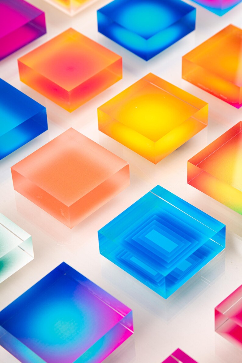 Puede incluir: Una serie de bloques cuadrados transl&uacute;cidos de colores en tonos azules, naranjas, amarillos y rosas. Los bloques est&aacute;n dispuestos sobre una superficie blanca, creando un dise&ntilde;o vibrante y moderno. Algunos bloques tienen un interior en capas.