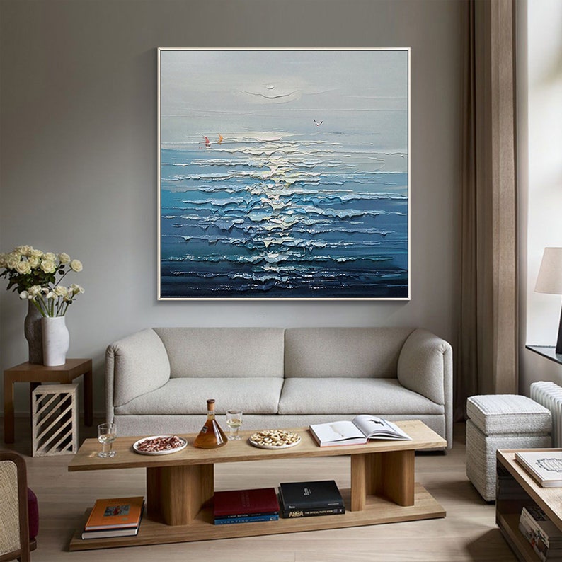 May include: A painting of a seascape with blue and white waves and a light blue sky. The painting is framed in a white frame and is hanging on a wall above a gray sofa.