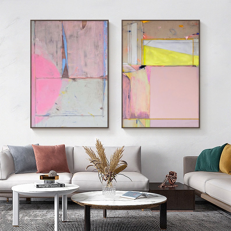 May include: Two framed abstract paintings in a modern style. The artwork uses pink, grey, yellow, and blue colours. The paintings are displayed above a white sofa with decorative cushions and a coffee table.