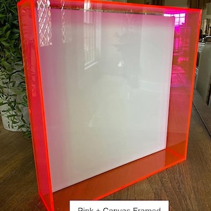 May include: A clear acrylic box frame with bright pink edges. The frame holds a white canvas. The text "Pink + Canvas Framed" is at the bottom of the frame.