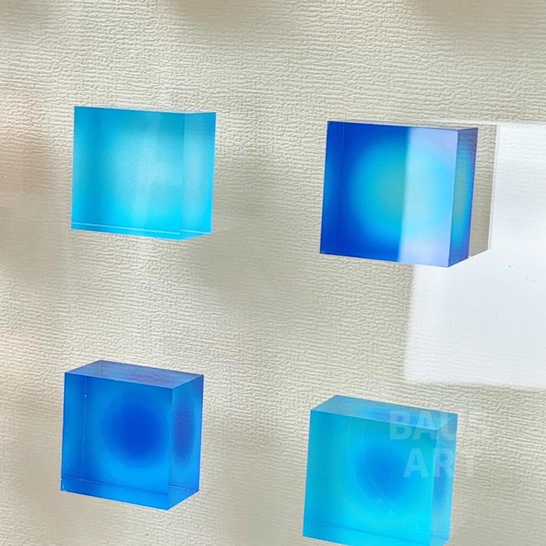 May include: Four square, translucent blue resin blocks with a white background. The blocks have a gradient of blue from light to dark.
