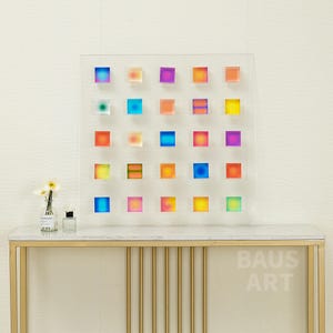May include: A wall art piece featuring a grid of 25 colorful squares with gradient patterns. The squares are arranged in a 5x5 grid on a clear acrylic background. The art piece is displayed on a white marble console table with gold metal legs. The table has the text "BAUS ART" printed on the front.