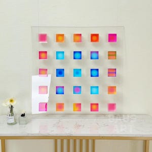 May include: A wall art piece with a grid of 25 colorful squares. The squares are made of clear acrylic and have a gradient of colors, including pink, orange, blue, and yellow.