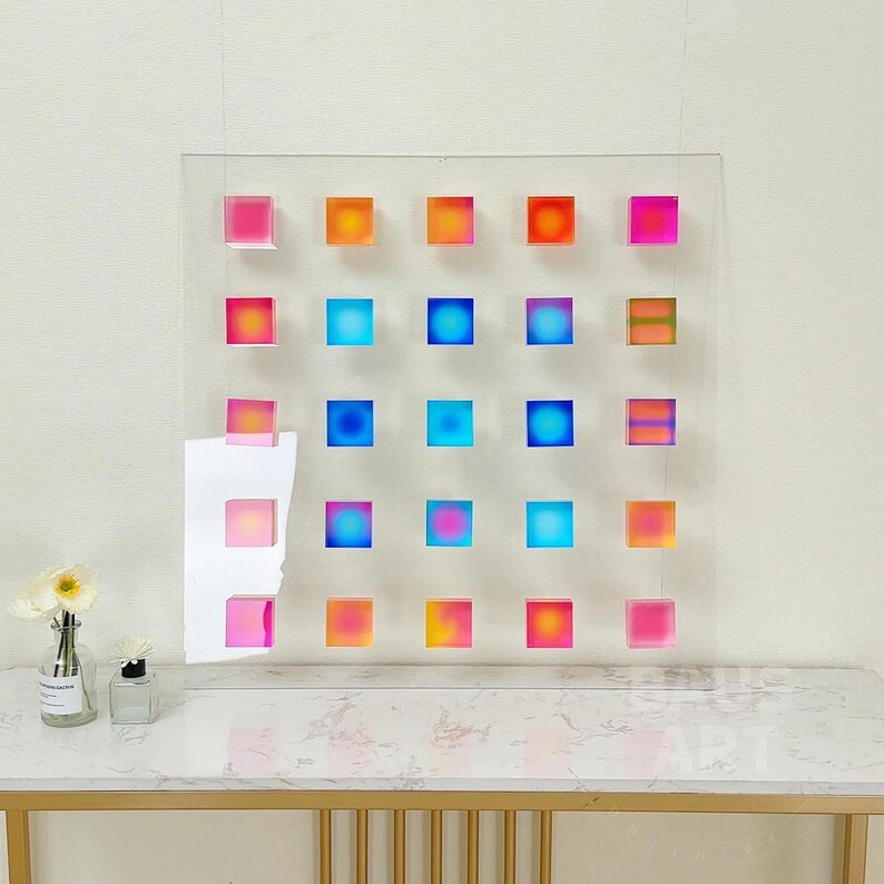 May include: A wall art piece with a grid of 25 colorful squares. The squares are made of clear acrylic and have a gradient of colors, including pink, orange, blue, and yellow.