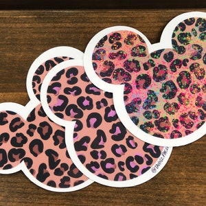 Op de afbeelding: Drie Mickey Mouse-hoofd stickers met een roze en zwarte luipaardprint en witte contouren. De bovenste sticker heeft een regenboog glitter-effect.