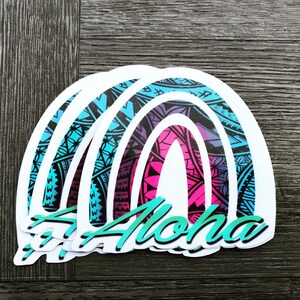 May include: A stack of four white stickers with a rainbow design and the word "Aloha" in teal. The rainbow is made up of blue, purple, pink, and teal colors and has a black and white patterned design.
