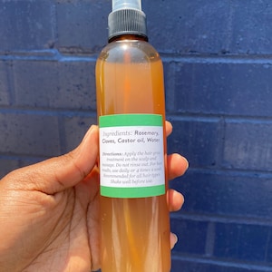 May include: A brown glass bottle with a clear spray nozzle filled with a light brown liquid. The bottle has a green label with the text "Ingredients: Rosemary, Cloves, Castor oil, Water. Directions: Apply the hair grow treatment on the scalp and massage. Do not rinse out. For best results, use daily or 4 times a week. Recommended for all hair types Shake well before use."