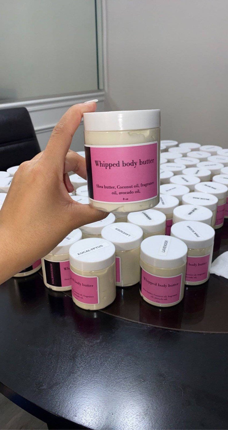 May include: A collection of white jars with pink labels containing whipped body butter. The label on the front jar reads "Whipped body butter" and lists the ingredients: Shea butter, Coconut oil, fragrance oil, avocado oil. The jars are arranged on a black table.