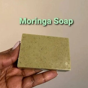 May include: A green bar of Moringa Soap, held in a hand.
