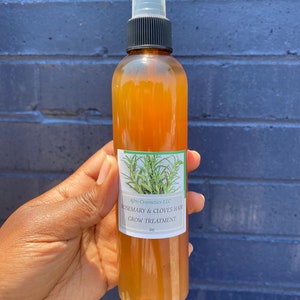 May include: A brown glass bottle with a black spray top and a white label. The label reads "Afro Cosmetics LLC Rosemary & Cloves Hair Grow Treatment 8oz".