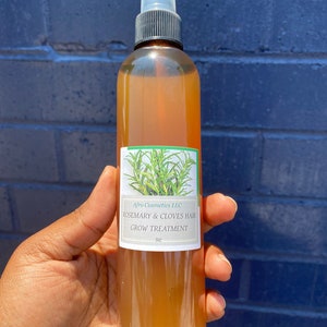 May include: A clear glass bottle with a brown cap and a white spray nozzle. The bottle is filled with a light brown liquid and has a white label that reads "Afro Cosmetics LLC Rosemary & Cloves Hair Grow Treatment 8oz".
