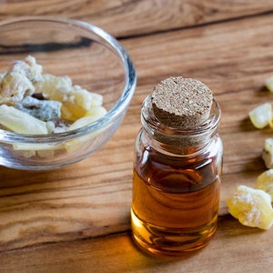 May include: A small glass bottle with a cork stopper filled with amber-colored liquid. The bottle is sitting on a wooden surface next to a small bowl of white and yellow resin pieces.