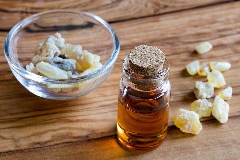 May include: A small glass bottle with a cork stopper filled with amber-colored liquid. The bottle is sitting on a wooden surface next to a small bowl of white and yellow resin pieces.