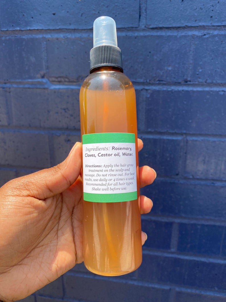 May include: A brown glass bottle with a clear spray nozzle filled with a light brown liquid. The bottle has a green label with the text "Ingredients: Rosemary, Cloves, Castor oil, Water. Directions: Apply the hair grow treatment on the scalp and massage. Do not rinse out. For best results, use daily or 4 times a week. Recommended for all hair types Shake well before use."