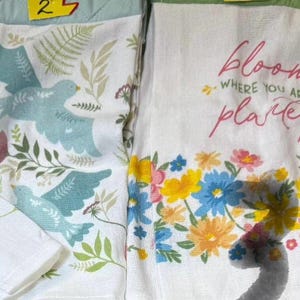 May include: White kitchen towels with floral and bird designs. One towel features blue birds and foliage. The other towel has the text "bloom where you are planted" with colorful flowers. A decorative and functional kitchen accessory.