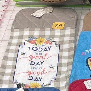 May include: A quilted oven mitt with a gray and white checkered pattern. The mitt features the text "TODAY is a good DAY for a good DAY" with floral and bee accents. A yellow tag with the number 24 is attached.
