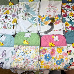 May include: Assortment of kitchen textiles, including oven mitts and dish towels. The towels feature various floral, butterfly, and bee designs. The oven mitts are quilted and come in green, pink, and blue. Some towels have text that reads "bloom where you are planted" and "enjoy the little things."
