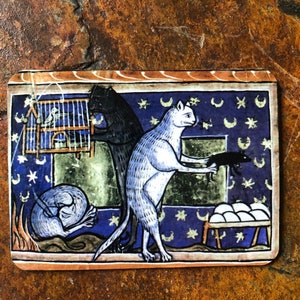 Op de afbeelding: Een middeleeuwse illustratie met drie katten. Een witte kat met een lange staart houdt een zwarte kat in zijn bek. Een zwarte kat houdt een vogelkooi vast met een vogel erin. De achtergrond is een blauw en gouden patroon met sterren en halve manen.